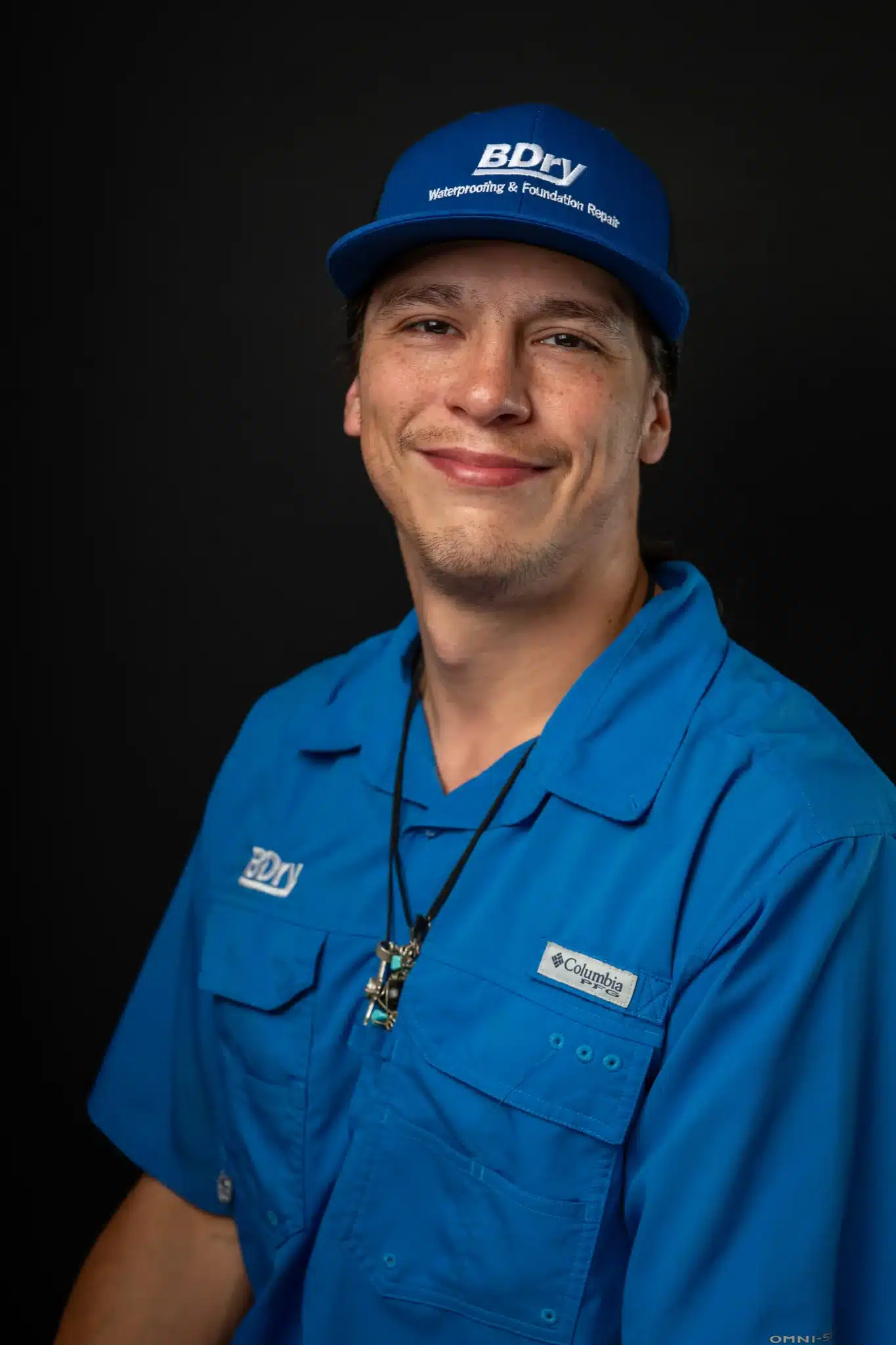 Man in blue uniform and cap with logo, black background, smiling.