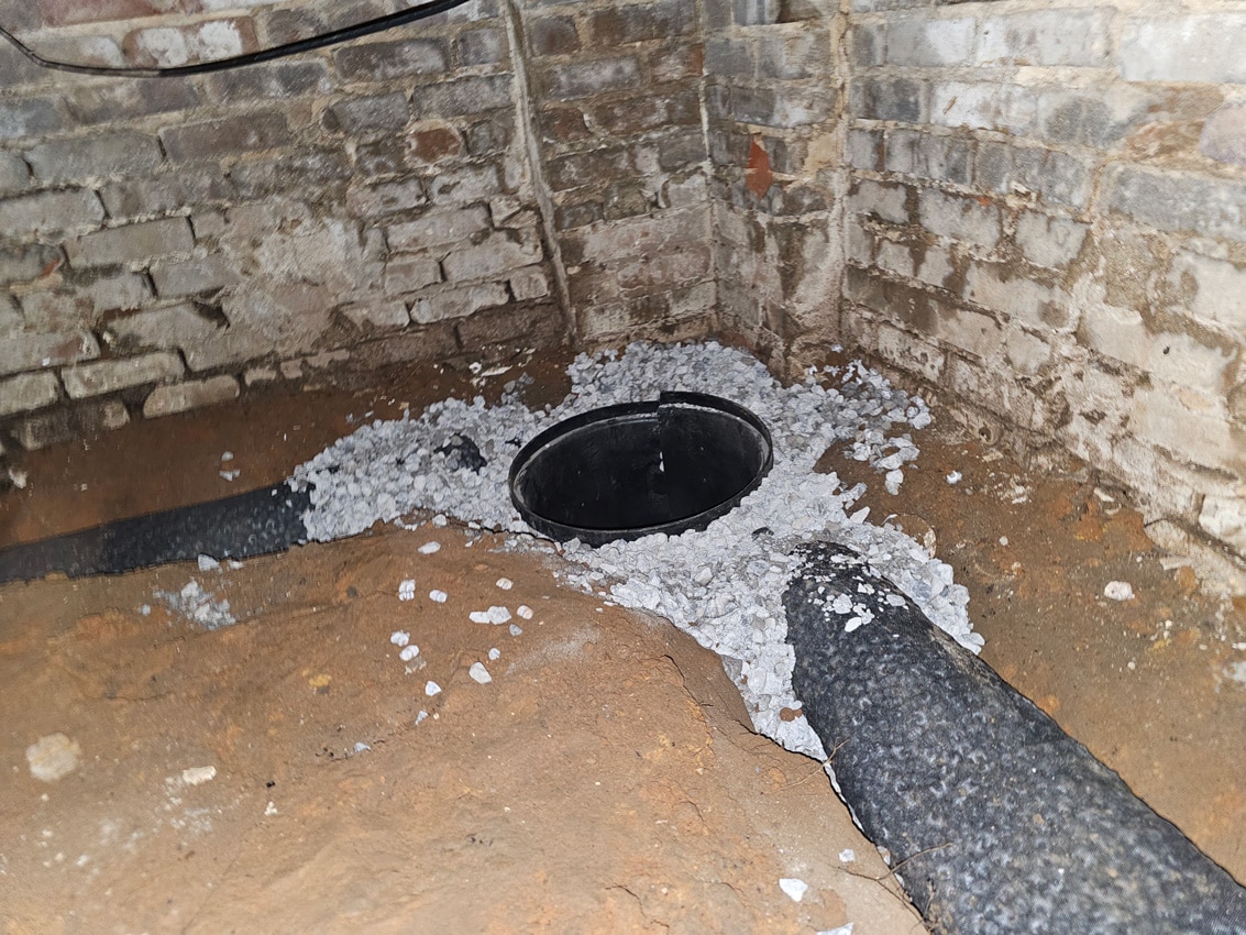 Sump pump installation with gravel and pipes in a basement corner with brick walls.