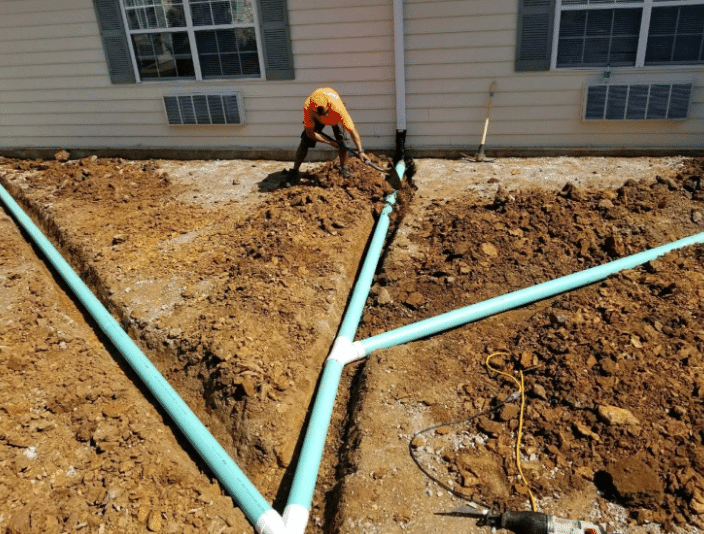 Worker installing drainage pipes in trench outside a building.