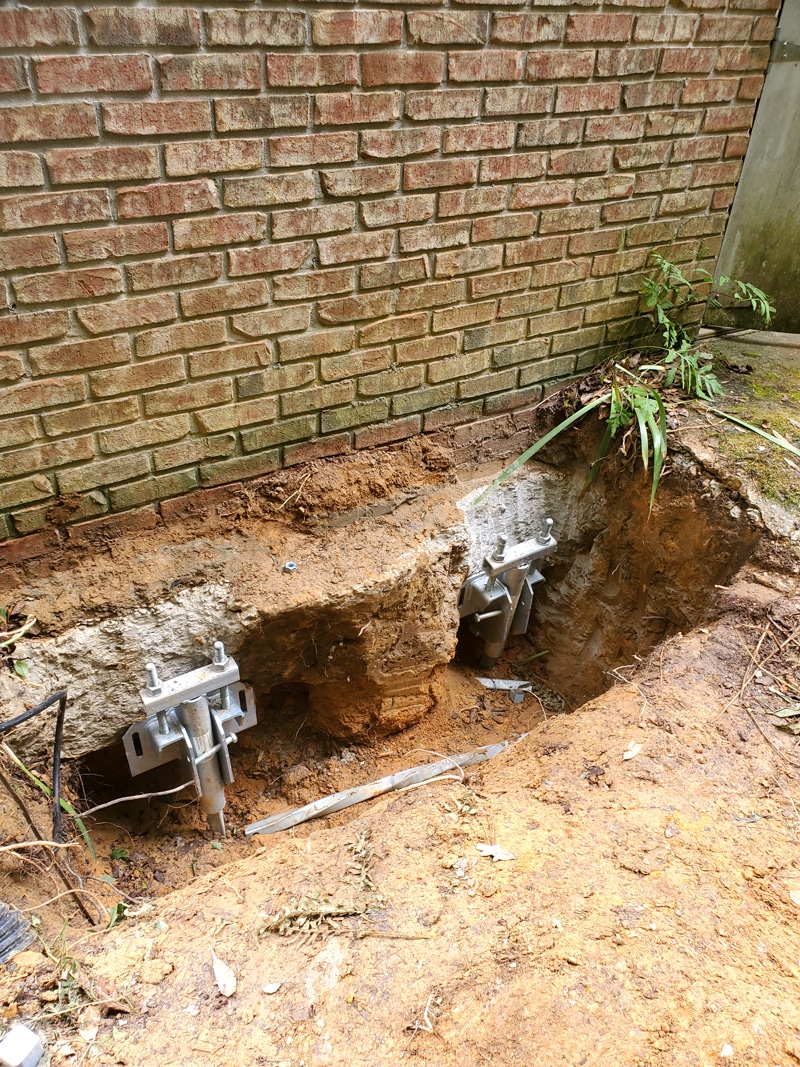 Brick wall with exposed foundation and helical piers in excavated soil for structural repair.