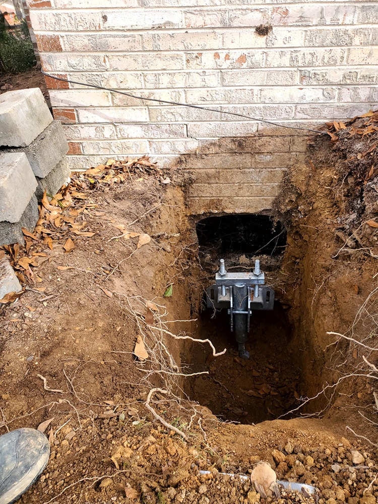 Foundation repair with a hydraulic jack near a brick wall, surrounded by soil and leaves.