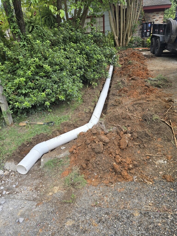 Trench with white drainage pipe installation beside a driveway and greenery.