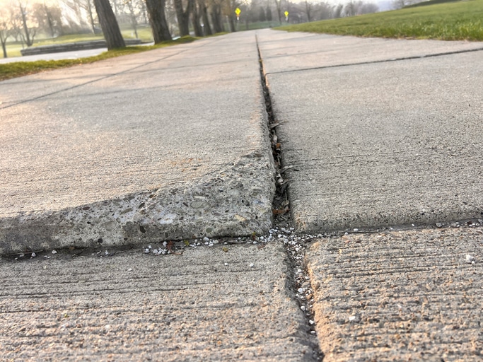 Cracked sidewalk with uneven surface, view along the pathway lined with grass and trees.