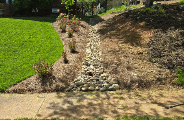 Dry creek bed in landscaped yard with rocks and plants for drainage and aesthetic appeal.