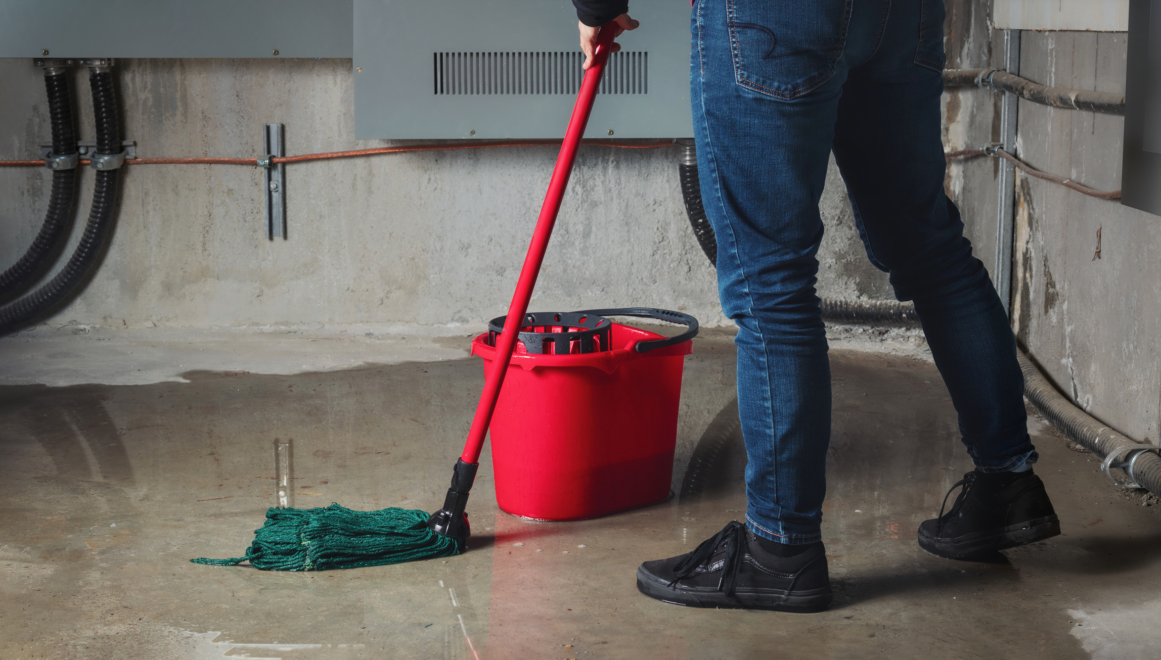 A woman mopping a flooded basement with water leaking through cracked concrete walls — a serious basement moisture problem.